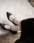 A person in a black skirt sits on a light stone surface, wearing Unified Sheer Bubble Socks and black Mary Jane shoes.