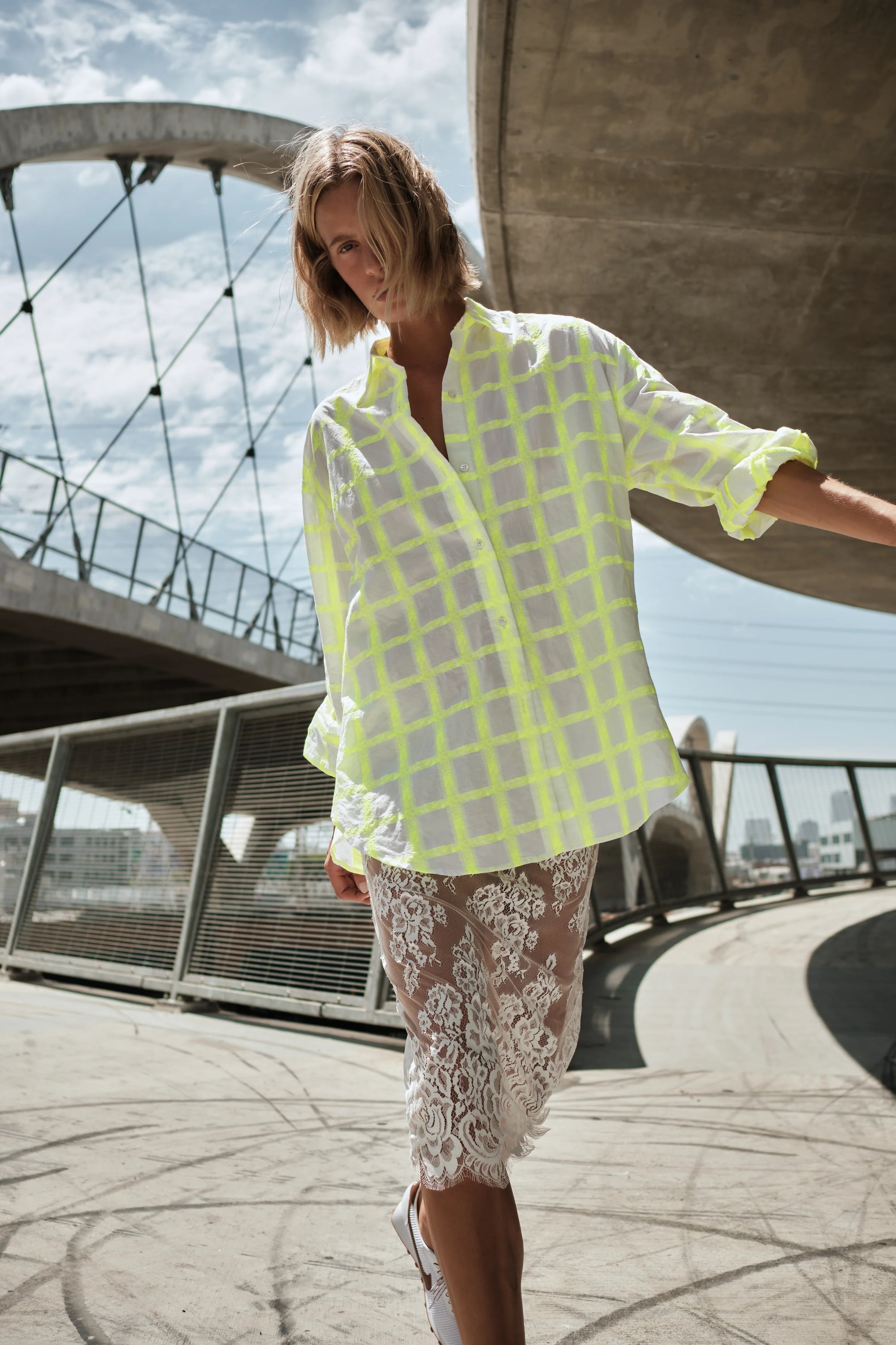 A person stands outdoors in Cissa’s The Victorian Shirt, a loose neon yellow checkered top, over a white lace pencil skirt with modern bridge architecture and a partly cloudy sky in the background.