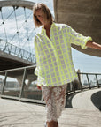 A woman poses outdoors under a partly cloudy sky, gracefully extending one arm while wearing The Victorian Shirt by Cissa—a loose, neon yellow checkered shirt—over a white lace pencil skirt with an adjustable waistband near a modern bridge.
