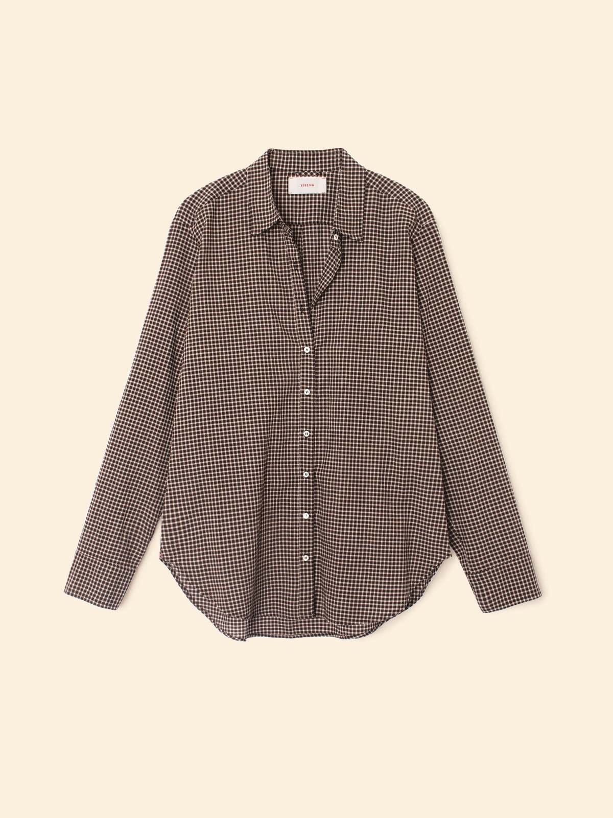 The Beau Shirt by Xirena is a menswear-inspired, long-sleeve button-up in black and white small checks, made from crisp cotton poplin with a collar and curved hem, shown against a plain beige background.