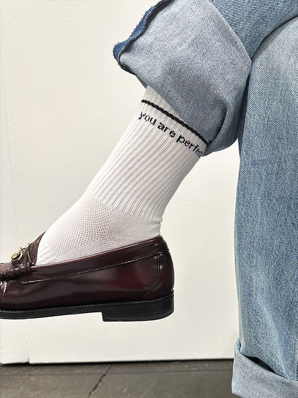 A person wearing light blue jeans and brown loafers has their leg crossed, revealing the Perfectwhitetee "You Are Perfect Sock" in white ribbed cotton with black text reading "you are perfect.