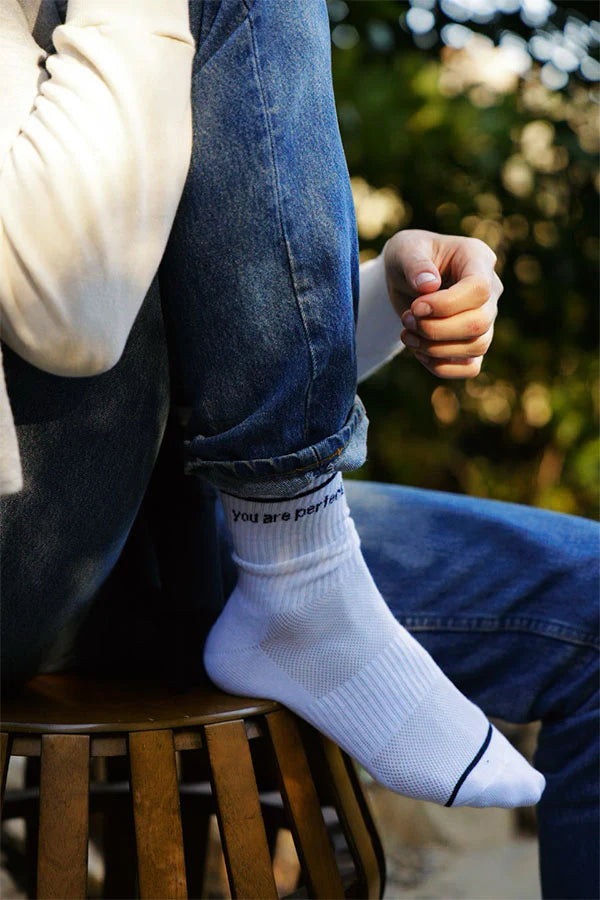 A person in jeans and a light sweater sits on a wooden stool, wearing Perfectwhitetee’s You Are Perfect Sock—vintage-inspired ribbed cotton socks with “you are perfect” near the top. Blurred greenery forms the backdrop.