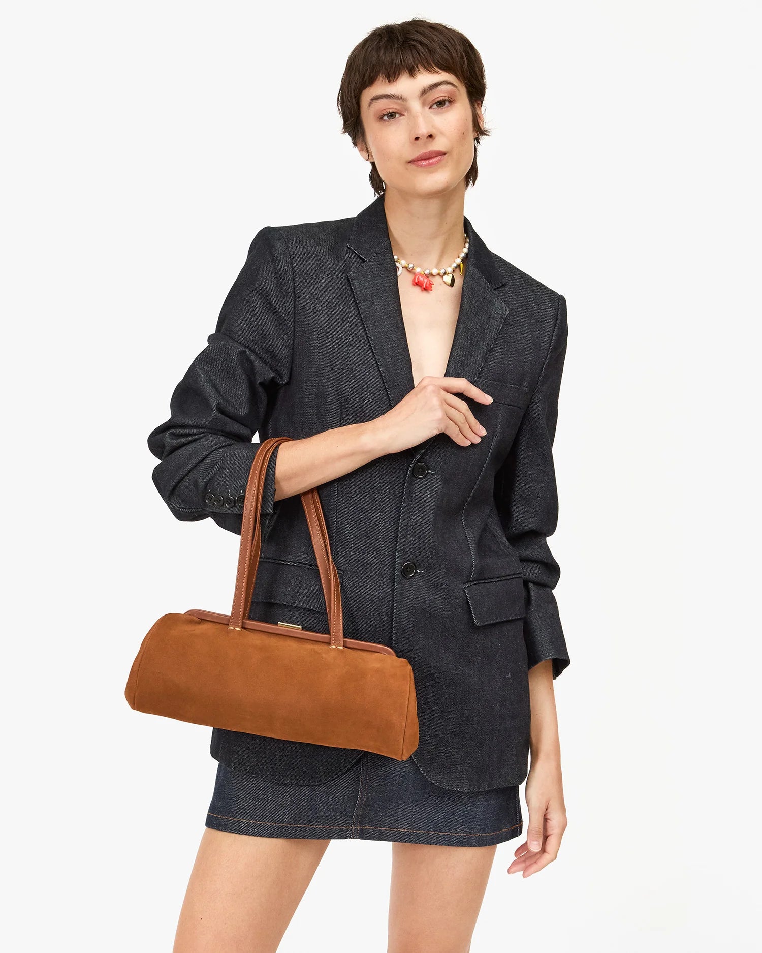 Wearing a dark denim blazer and matching skirt, a person with short hair holds the Clare Vivier Yvette Smooth Nubuck shoulder bag. They accessorize with a colorful beaded necklace, all set against a plain light background.