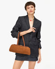 Wearing a dark denim blazer and matching skirt, a person with short hair holds the Clare Vivier Yvette Smooth Nubuck shoulder bag. They accessorize with a colorful beaded necklace, all set against a plain light background.