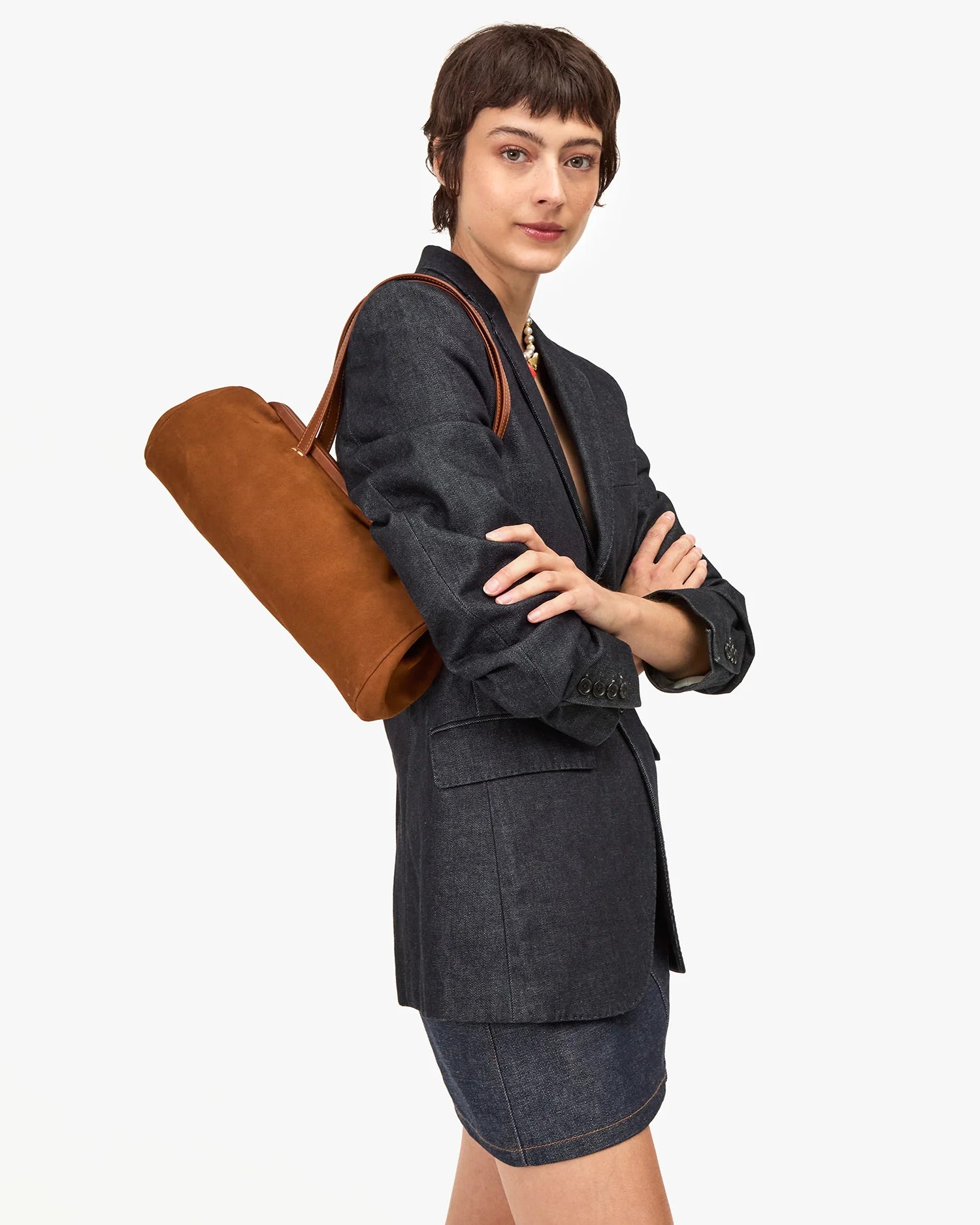 A woman with short brown hair in a dark blazer and denim skirt stands confidently with folded arms, modeling the Clare Vivier Yvette Smooth Nubuck shoulder bag against a plain white background.