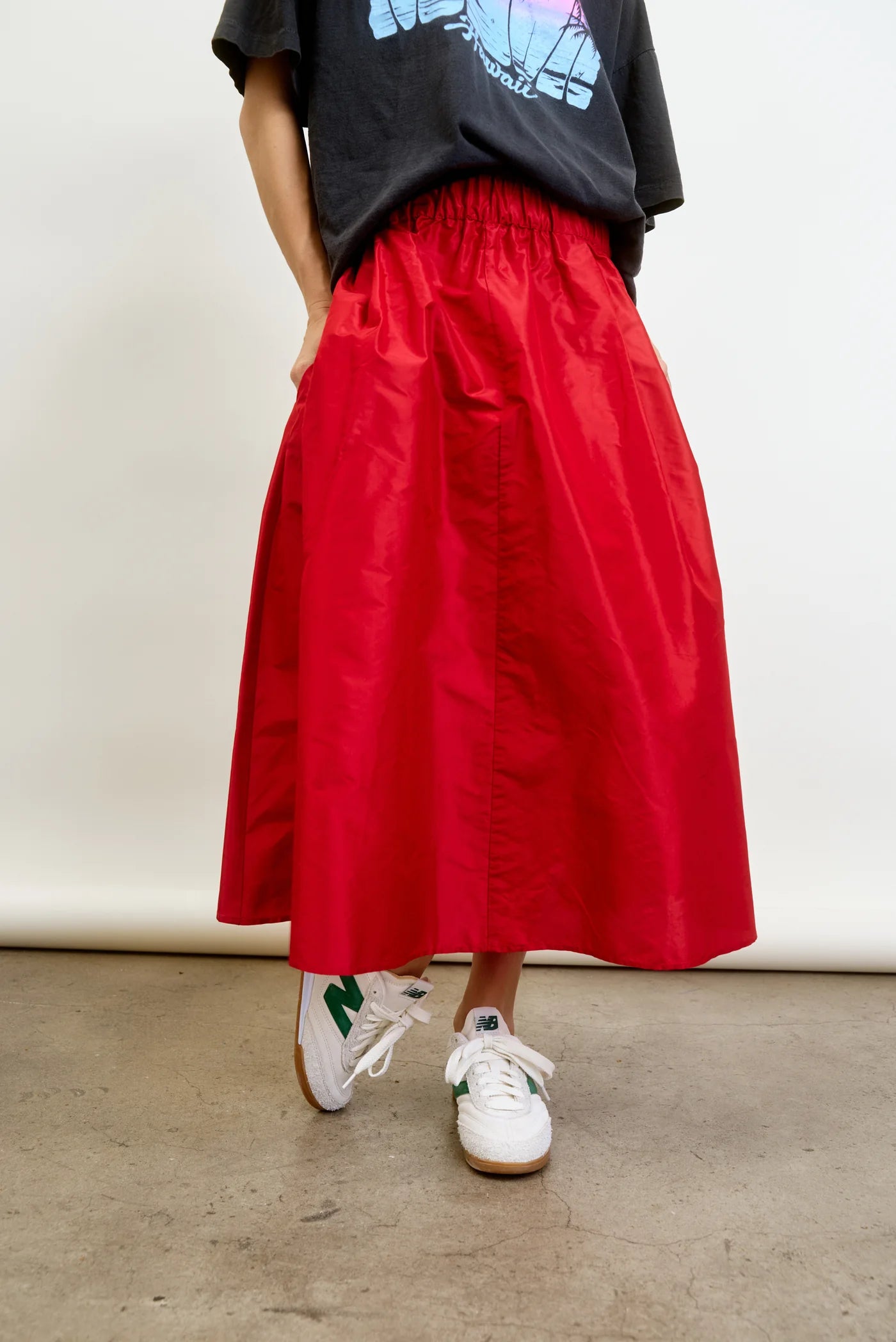 A person in a black graphic T-shirt and the Aquarius Cocktail LE SKIRT Silk Taffeta, a long red 40&#39;s inspired full-flared skirt with pockets and an elastic waistband, paired with white sneakers featuring green accents, stands against a white backdrop on a concrete floor.