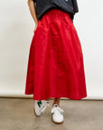 A person in a black graphic T-shirt and the Aquarius Cocktail LE SKIRT Silk Taffeta, a long red 40's inspired full-flared skirt with pockets and an elastic waistband, paired with white sneakers featuring green accents, stands against a white backdrop on a concrete floor.