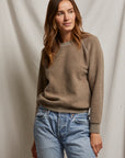 A person with long hair, draped in the Ziggy Inside Out LS Crewneck Sweatshirt from Perfectwhitetee and light blue jeans, stands against a neutral fabric backdrop. Featuring exposed seam detailing, they strike a relaxed pose with one hand in their pocket and a gentle, thoughtful expression.