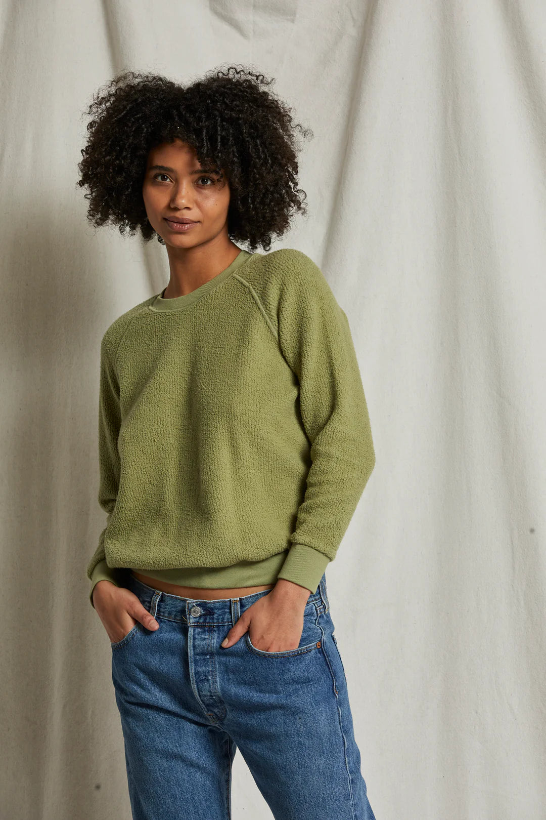 A person with short curly hair is wearing a green Ziggy Reverse Fleece LS Crew Sweatshirt from Perfectwhitetee, featuring exposed seam detailing, and blue jeans. They are standing in front of a plain light-colored backdrop with one hand in their pocket and the other relaxed by their side. The expression on their face appears calm.