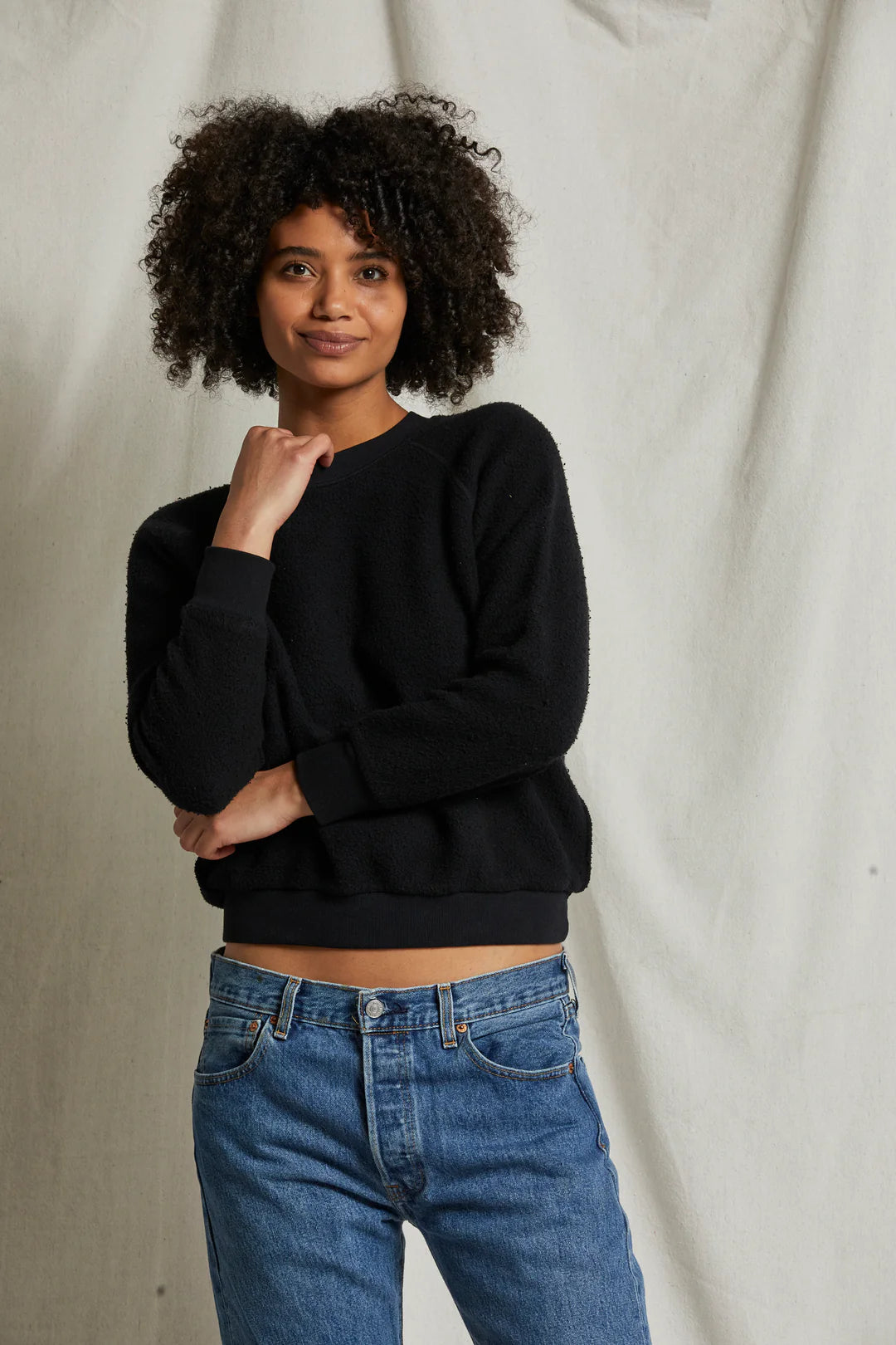 A person with curly hair stands in front of a neutral backdrop, wearing a cozy Perfectwhitetee Ziggy Inside Out LS Crewneck Sweatshirt in black and blue jeans. They have their right arm crossed and are gently touching their chin with their left hand, smiling softly at the camera.