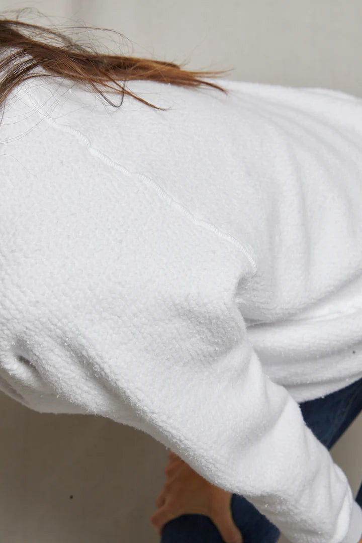 A close-up of a person wearing the Perfectwhitetee Ziggy Reverse LS Sweatshirt in textured white cotton fleece and blue jeans, leaning forward slightly. Only their upper body, arm, and hair are visible.