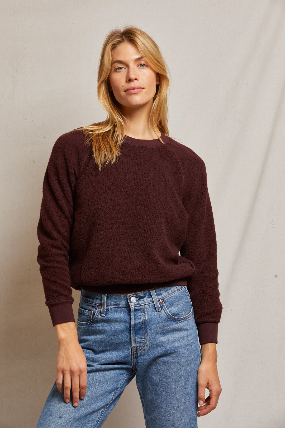 A person with long blonde hair is wearing the Ziggy Inside Out LS Crewneck Sweatshirt by Perfectwhitetee, crafted from buttery soft fleece in a dark brown shade, paired with blue jeans. They are standing against a plain beige background, looking confidently at the camera, embodying a cozy essential.