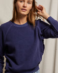 A person wearing the Ziggy Inside Out Longsleeve Crewneck Sweatshirt by Perfectwhitetee, a navy blue textured piece with exposed seam detailing, paired with light blue jeans stands against a neutral backdrop. They have a calm expression and are adjusting their hair with one hand.