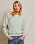 A person with long blonde hair is wearing a light green Ziggy Inside Out Longsleeve Crewneck Sweatshirt by Perfectwhitetee and blue jeans. They are smiling slightly with their hands in their pockets, standing against a plain beige background.