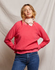 A person confidently poses against a neutral fabric backdrop, wearing the Ziggy Reverse Fleece LS Crew Sweatshirt by Perfectwhitetee in vibrant pink along with blue jeans and a light pink scarf. Their shoulder-length hair complements their cozy essential style.