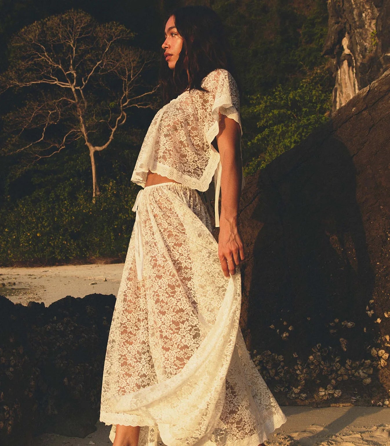 A woman stands on a sunlit beach, wearing the DOEN Zinnia Skirt—a sheer white lace two-piece with an asymmetrical hem and short sleeves—surrounded by rocks, greenery, and a bare tree in the background.