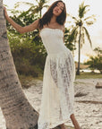 Wearing the DOEN Zinnia Skirt, a woman leans against a palm tree on a sandy beach, with sunlight and palms in the background.