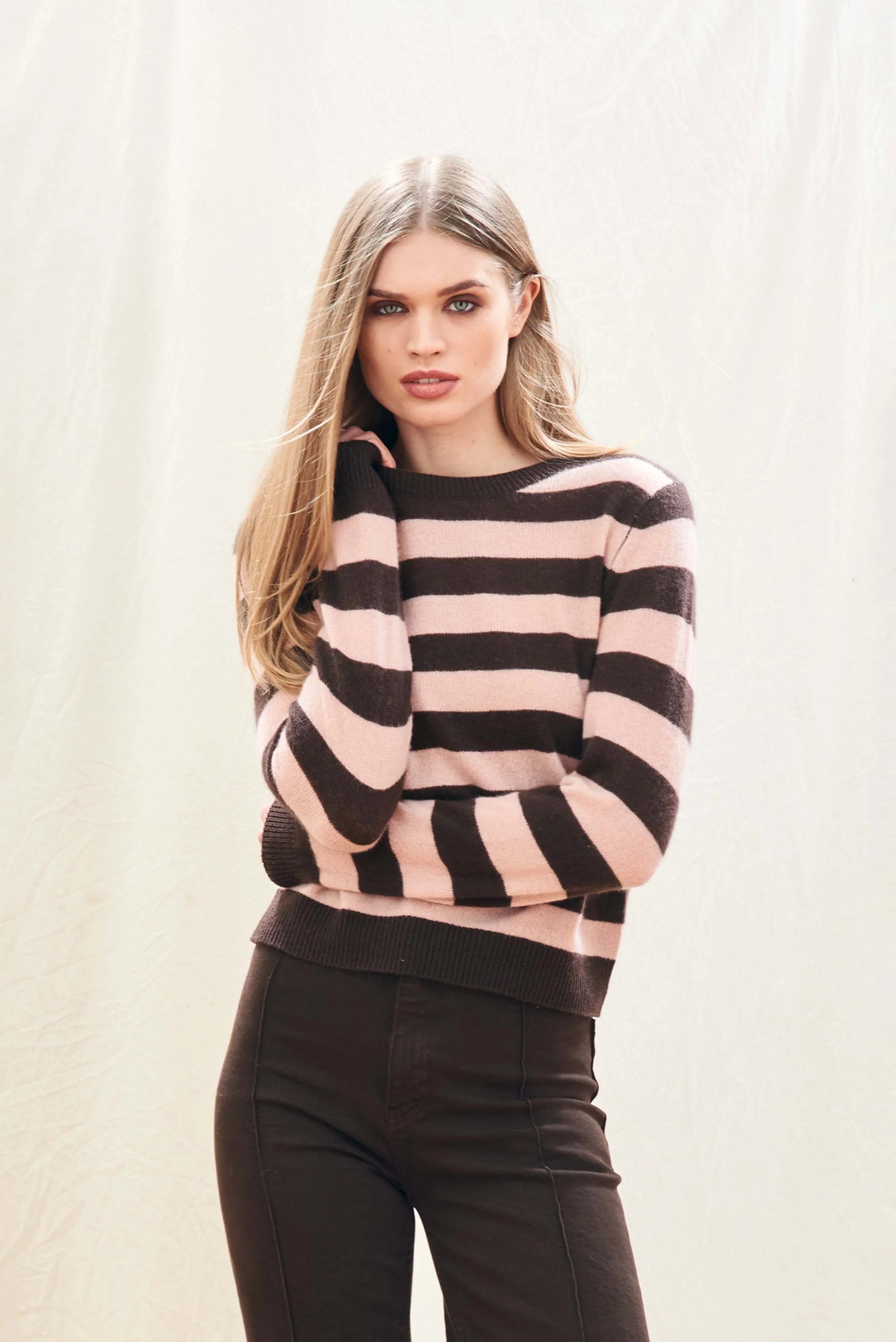 A person with long, straight blonde hair wears the Jumper 1 2 3 4/CR2 Cashmere Stripe Crew in pink and brown with black pants, standing against a plain, light background and touching their hair.