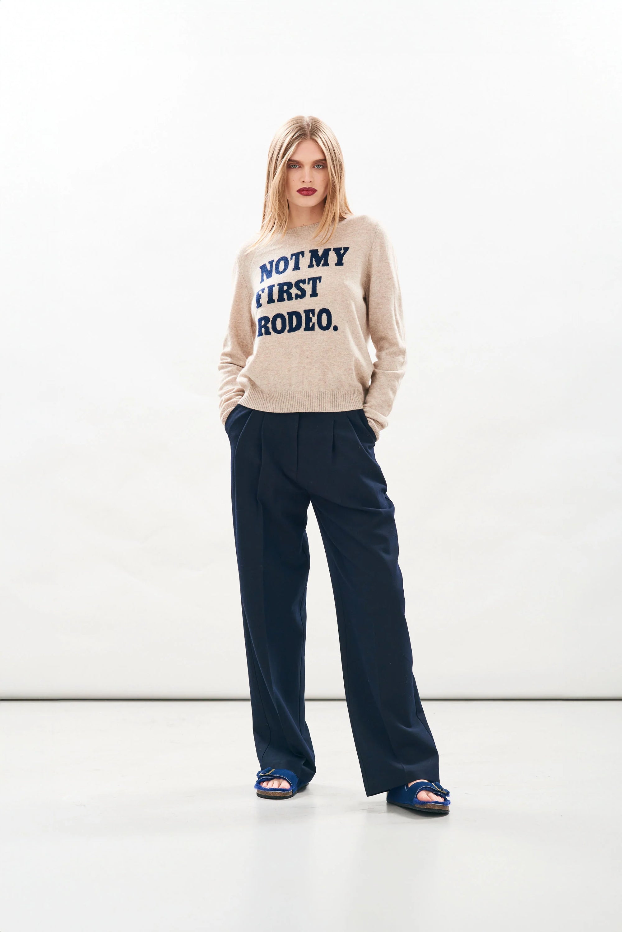 A woman poses confidently in the Rodeo Crew sweater by Jumper 1 2 3 4/CR2, featuring blue "NOT MY FIRST RODEO" text, paired with navy wide-leg trousers and blue sandals against a plain white background. Her hands are in her pockets.