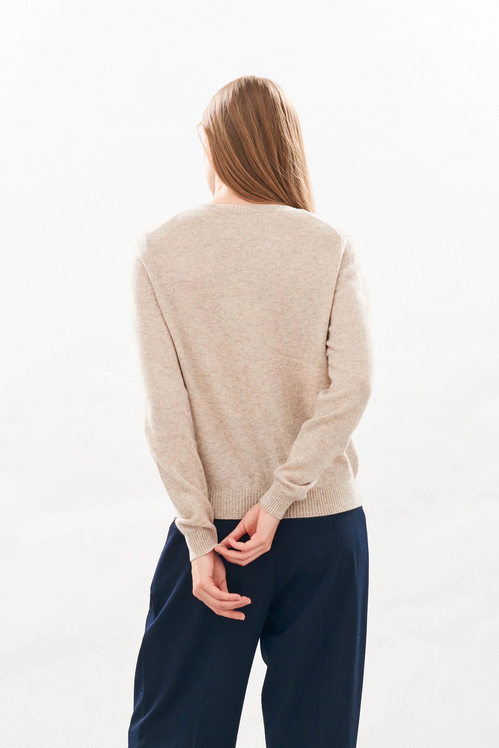 A person with long, straight hair stands facing away against a plain background, wearing the beige Rodeo Crew organic jumper by Jumper 1 2 3 4/CR2 and dark pants, hands behind their back, fingers loosely intertwined.