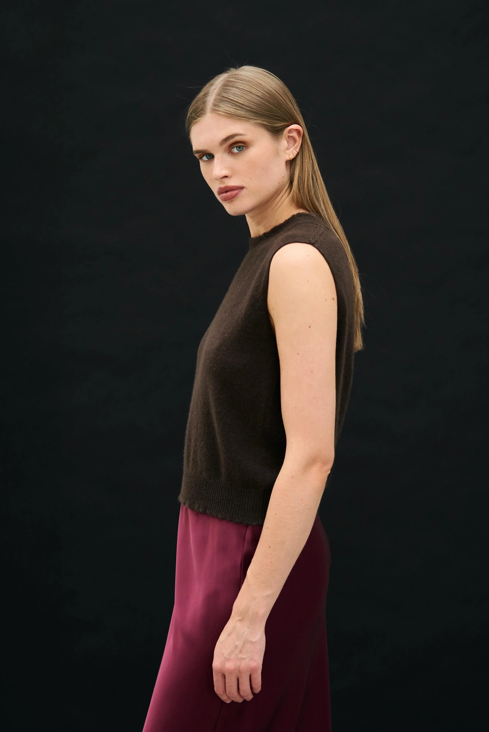 A woman with straight, light brown hair stands sideways against a plain black background, wearing the Jumper 1 2 3 4/CR2 Distressed Tank and maroon pants, calmly looking towards the camera.