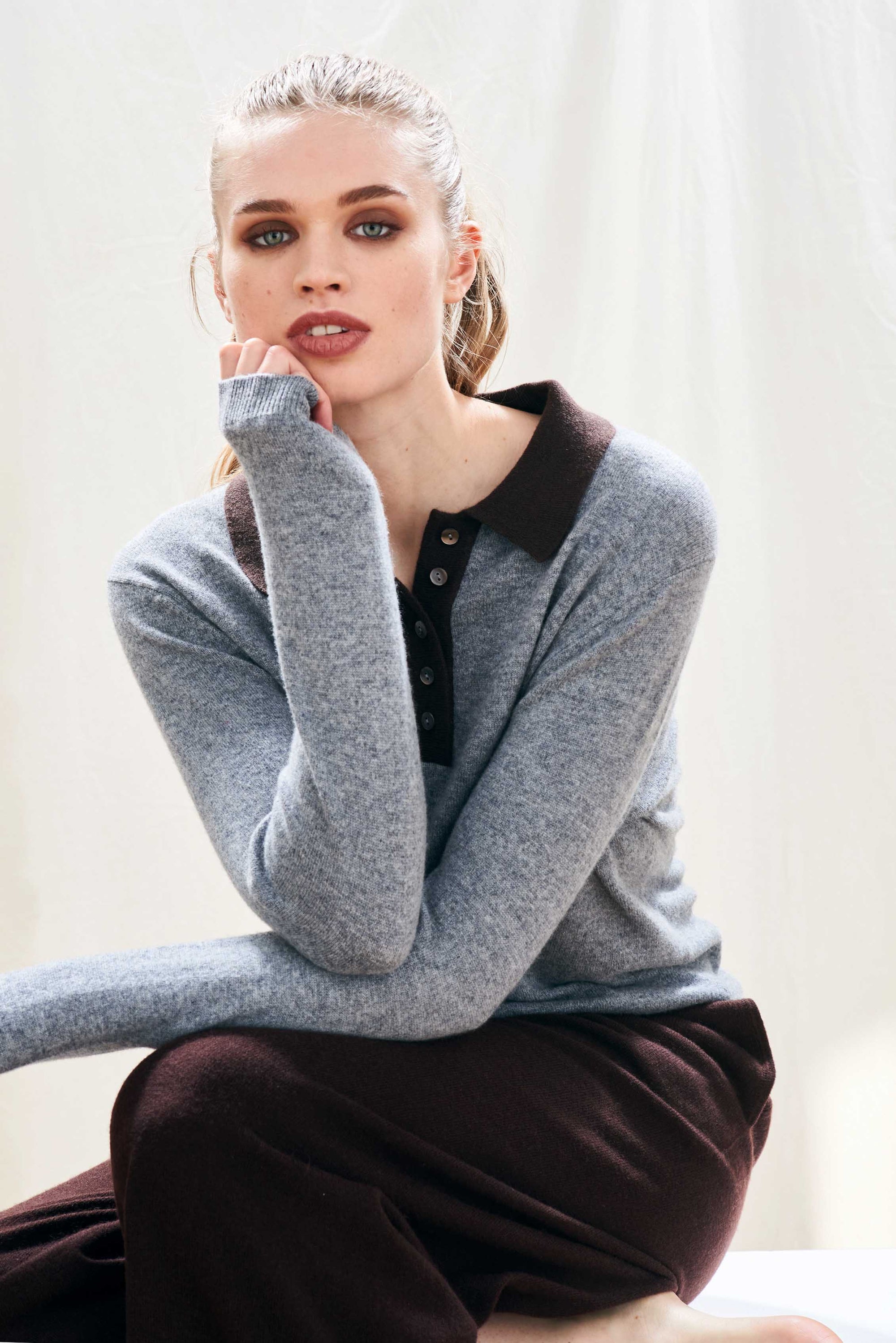 A woman with light hair pulled back wears a gray Jumper 1 2 3 4/CR2 Contrast Polo with a dark collar and brown pants, sitting with one arm bent and looking directly at the camera against a light background.
