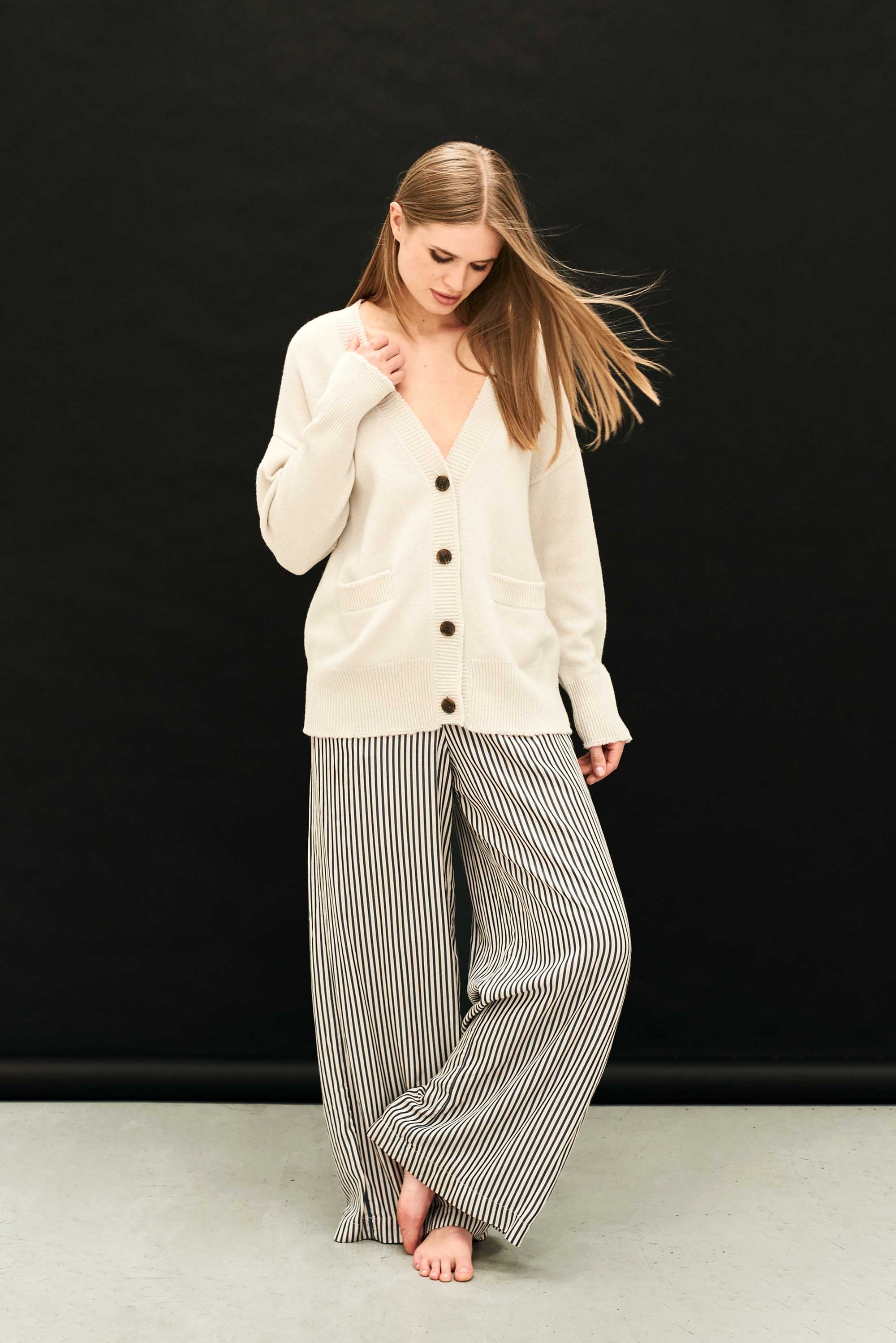 A woman stands barefoot against a black background, wearing the cream Boyfriend Heavy Cardigan by Jumper 1 2 3 4/CR2 and loose, black-and-white vertically striped pants. She looks down, her hair falling over one shoulder.