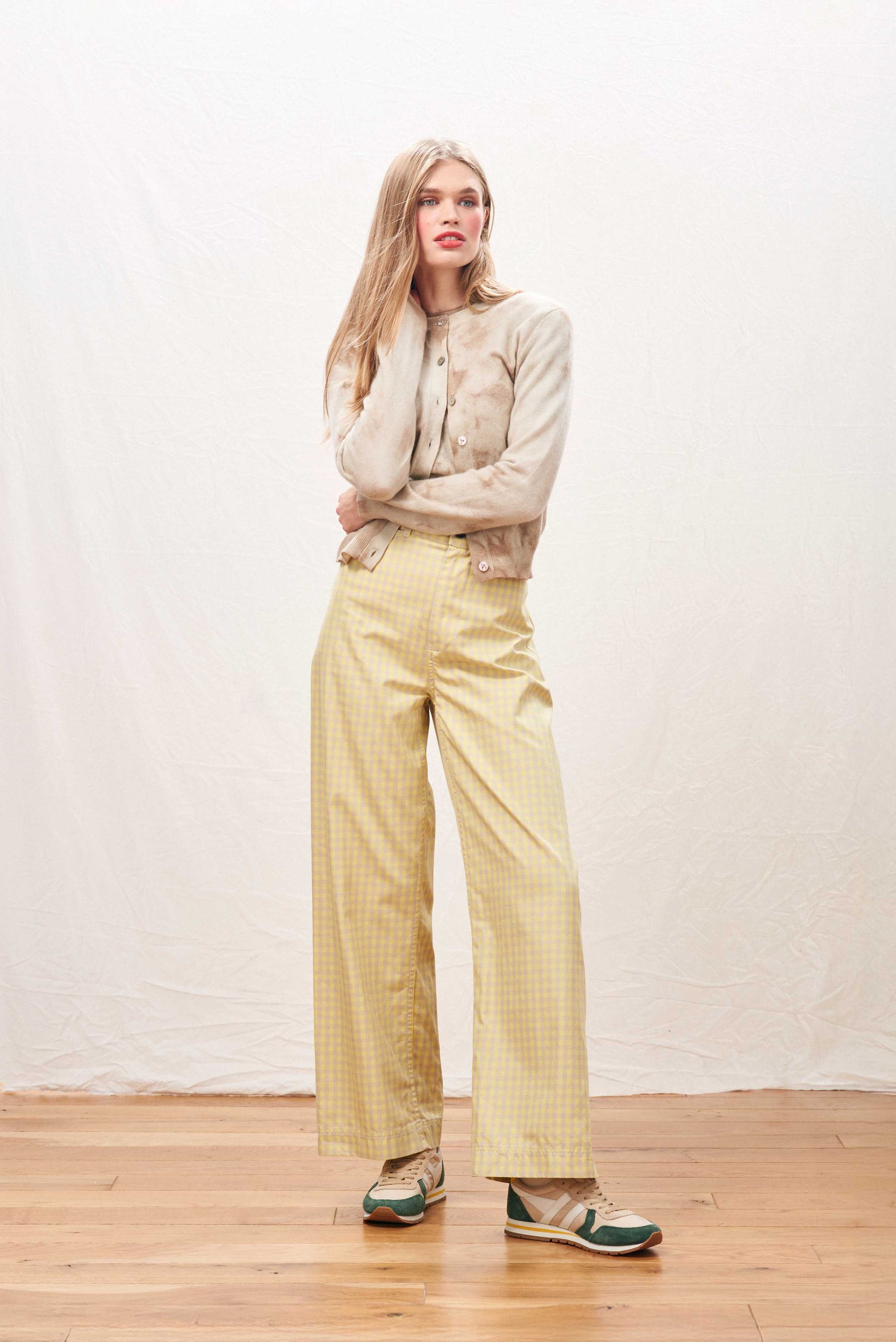 A woman stands indoors on a wooden floor, wearing the Batik Cardigan by Jumper 1 2 3 4/CR2, yellow checkered wide-leg pants, and light green sneakers, with a white backdrop behind her.