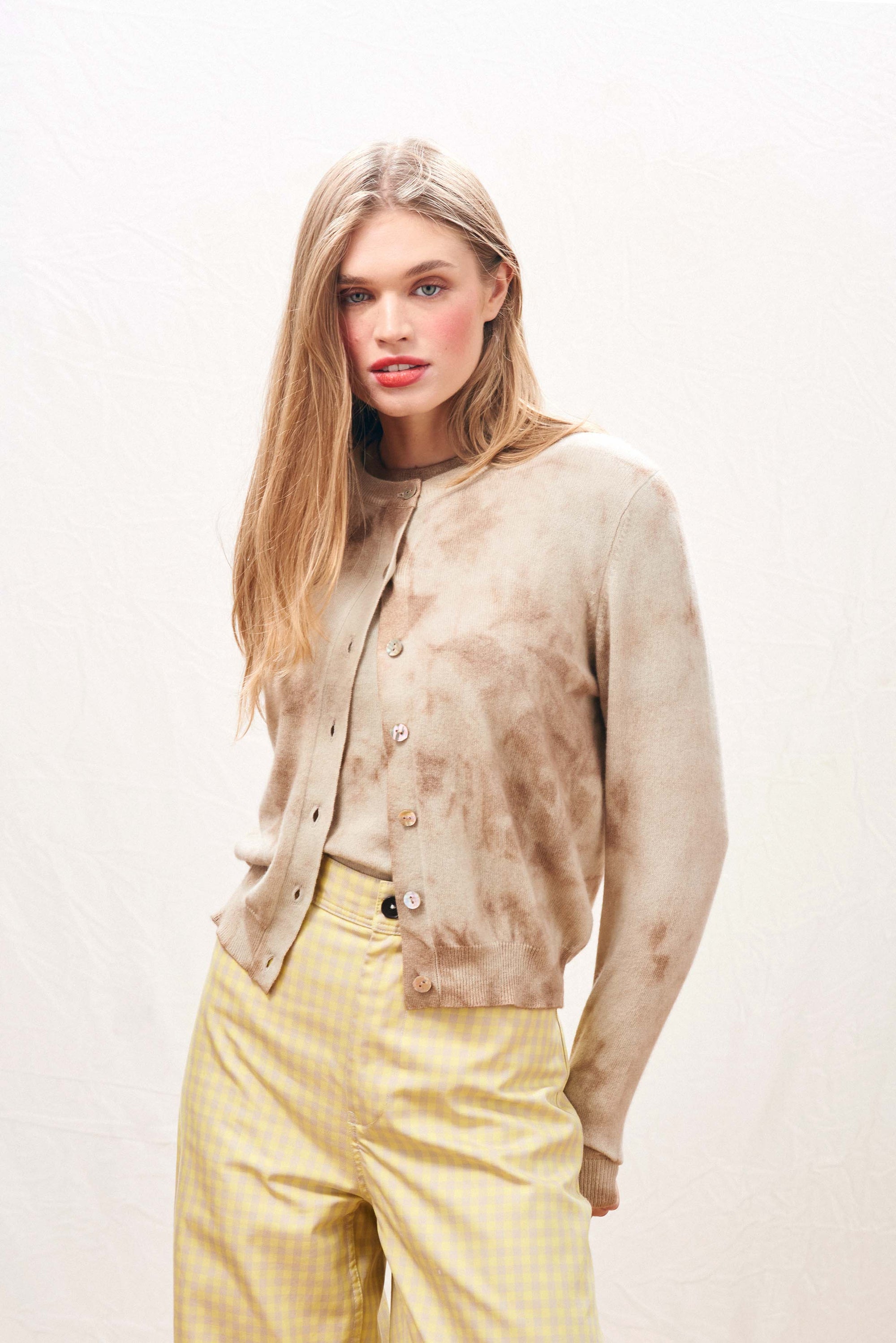 A woman with long blonde hair wears the Batik Cardigan by Jumper 1 2 3 4/CR2 over a matching top and light yellow gingham pants, standing against a light textured background and looking slightly to the side.