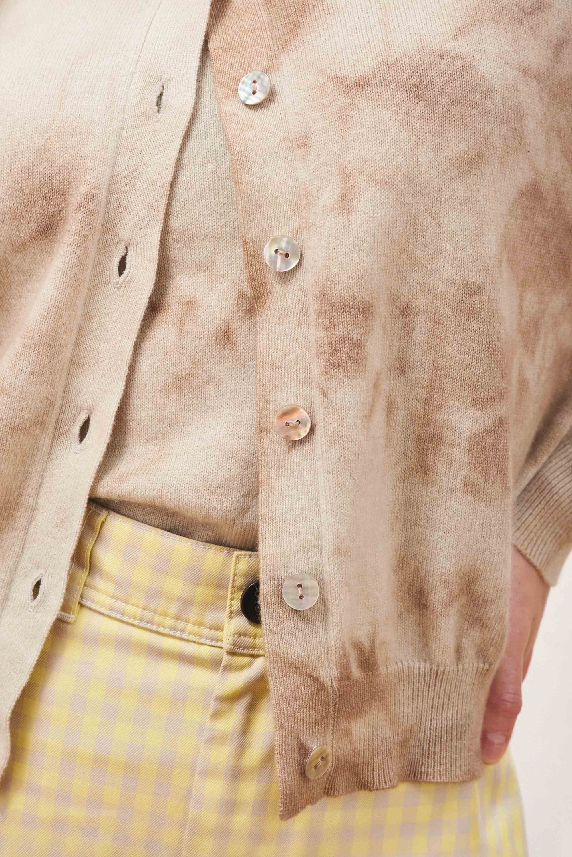 A close-up of a person wearing the Batik Cardigan by Jumper 1 2 3 4/CR2 over a matching top, styled with high-waisted yellow and white gingham pants, with their hand resting on their hip.