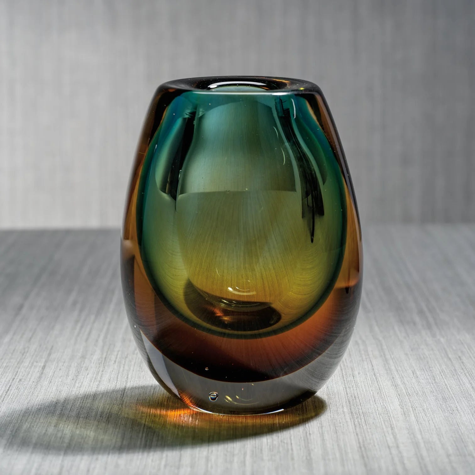 The Zodax Crystal Bloom Vase, featuring a smooth, glossy finish and a small opening, displays green, amber, and brown gradient hues. It sits on a light gray textured surface against a vertically striped gray background.