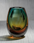 The Zodax Crystal Bloom Vase, featuring a smooth, glossy finish and a small opening, displays green, amber, and brown gradient hues. It sits on a light gray textured surface against a vertically striped gray background.