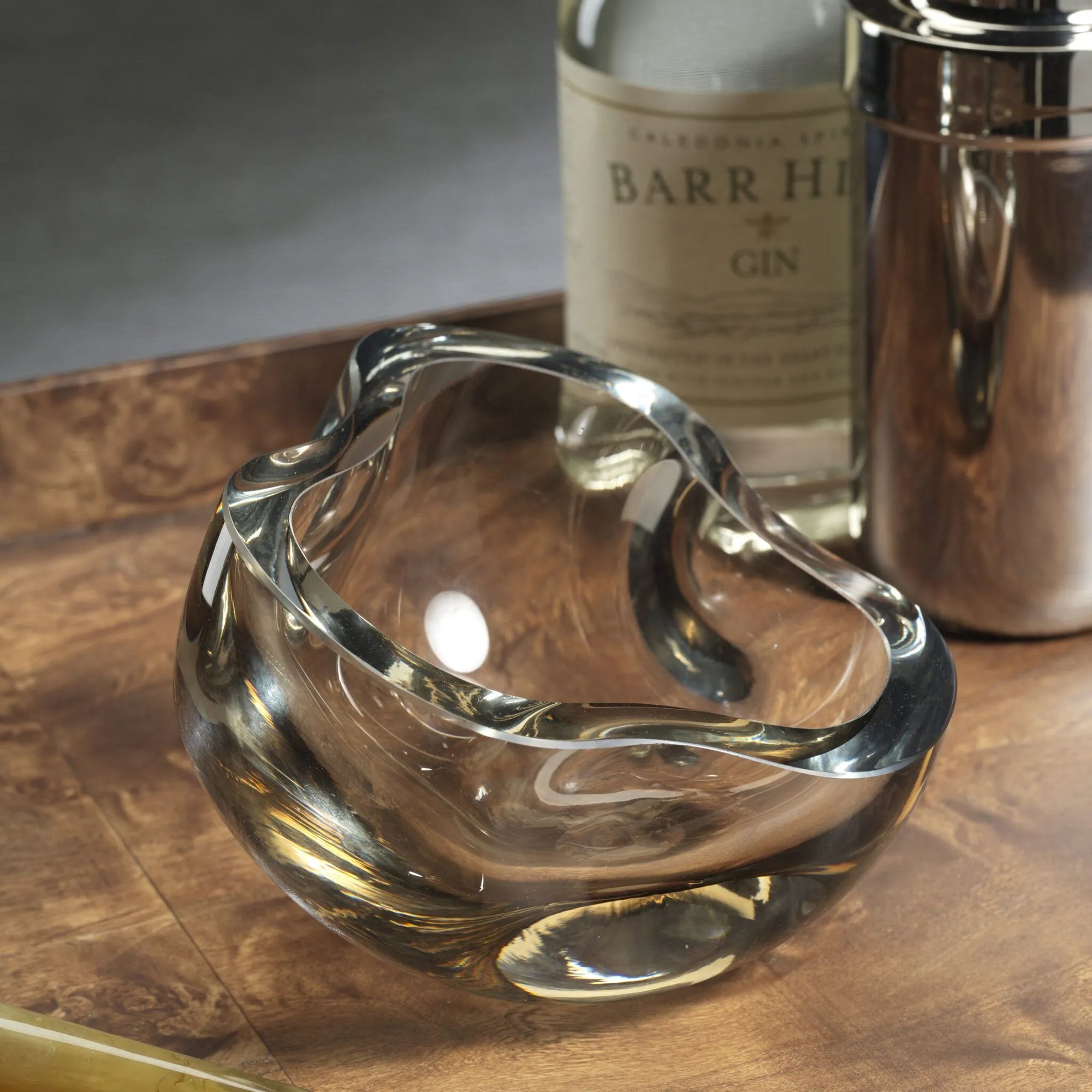 The Zodax CH-7877 Viento Glass Bowl, with its abstract organic shape, sits on a wooden tray beside Barr Hill Gin and a metal shaker, creating striking sculptural home decor for any space.