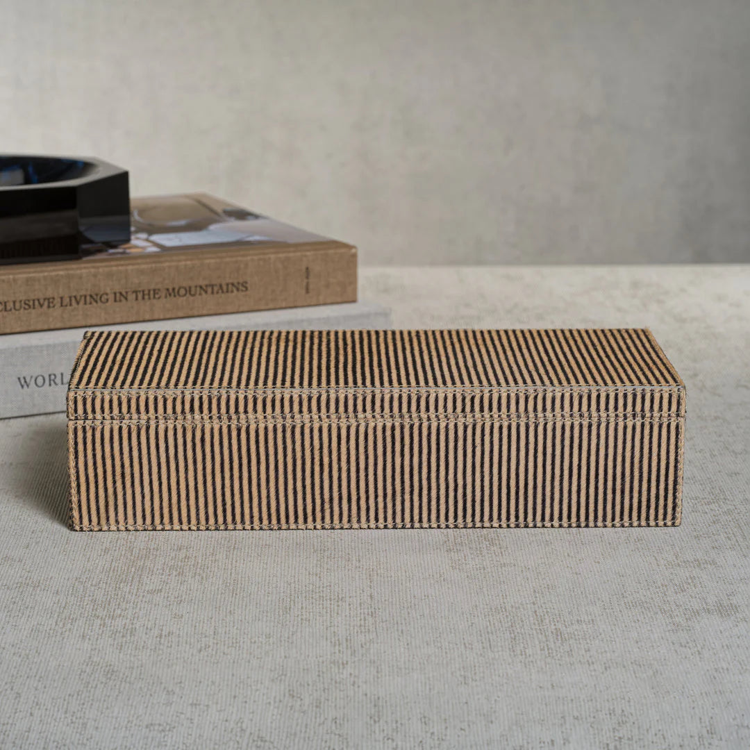 The Zodax Hair-On-Hide Leather Box, with elegant brown and black stripes, rests on a textured surface. Behind it, two stacked books—including "EXCLUSIVE LIVING IN THE MOUNTAINS"—enhance the sophisticated atmosphere.