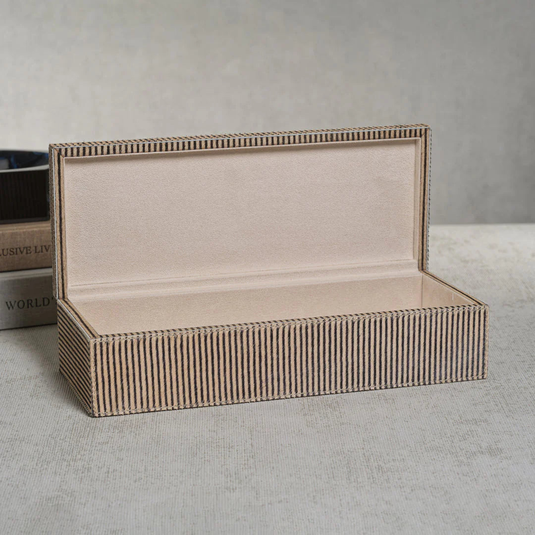 The Zodax Hair-On-Hide Leather Box features a rectangular, beige striped exterior and an open lid that reveals a soft, light interior—an elegant storage piece resting on a textured surface with a softly blurred background.