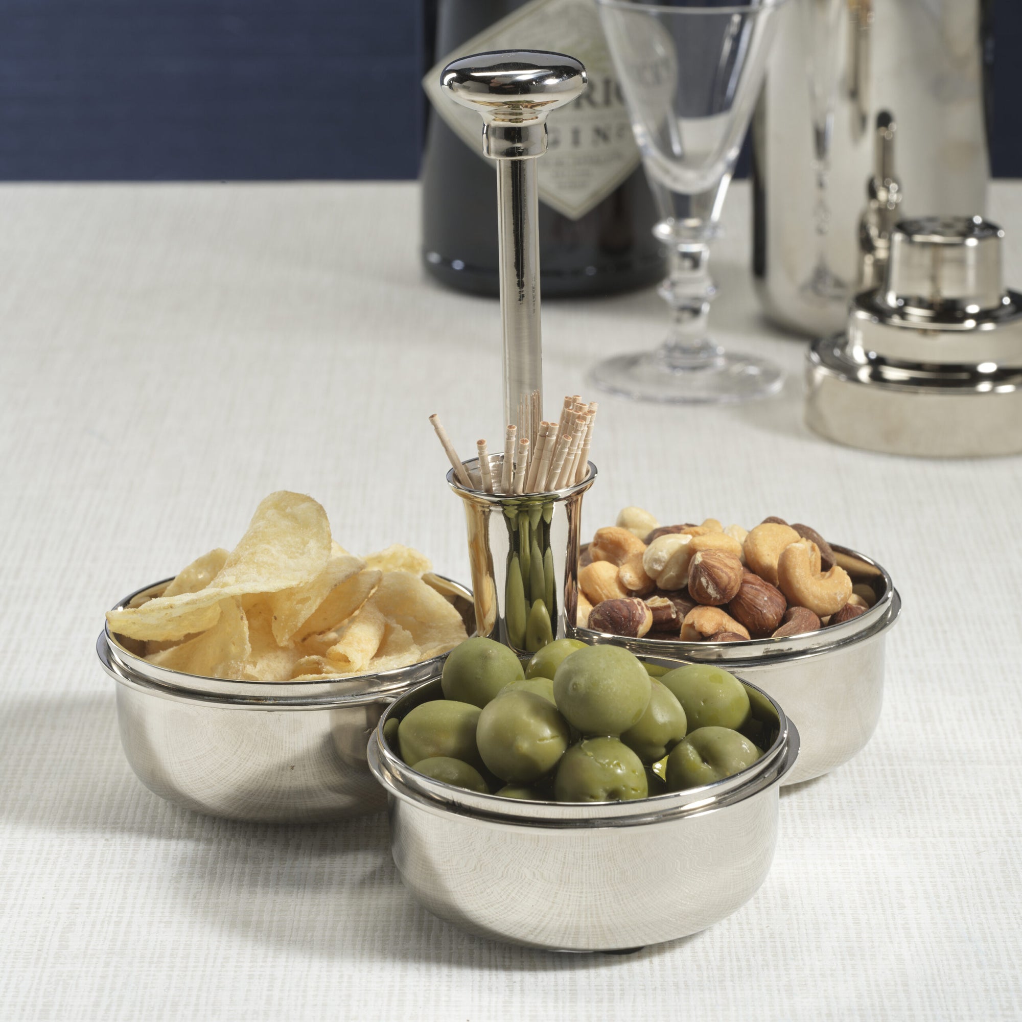 The Zodax Condiment Trio in polished nickel holds potato chips, green olives, and mixed nuts with a central cocktail pick holder. A cocktail shaker, martini glass, and gin bottle sit on a light tablecloth in the background.