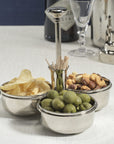 The Zodax Condiment Trio in polished nickel holds potato chips, green olives, and mixed nuts with a central cocktail pick holder. A cocktail shaker, martini glass, and gin bottle sit on a light tablecloth in the background.