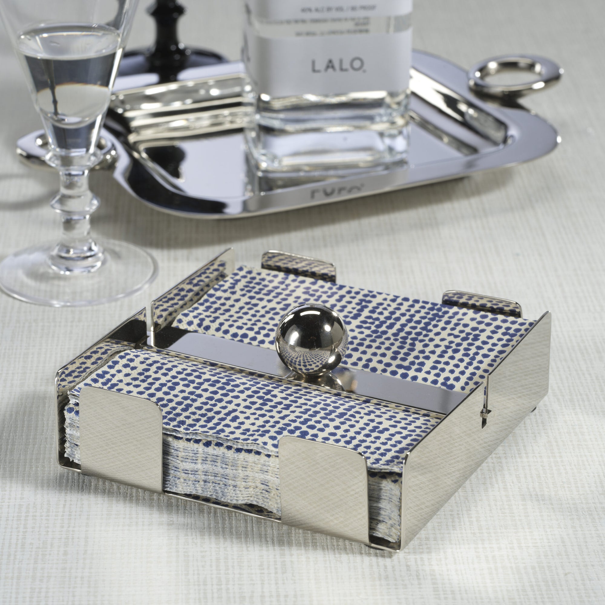 The Zodax Cocktail Napkin Holder Nickel, featuring a spherical weight, keeps blue and white napkins secure on your table. Perfect as a bar accessory, it sits elegantly beside a glass of water, silver tray, and decanter.