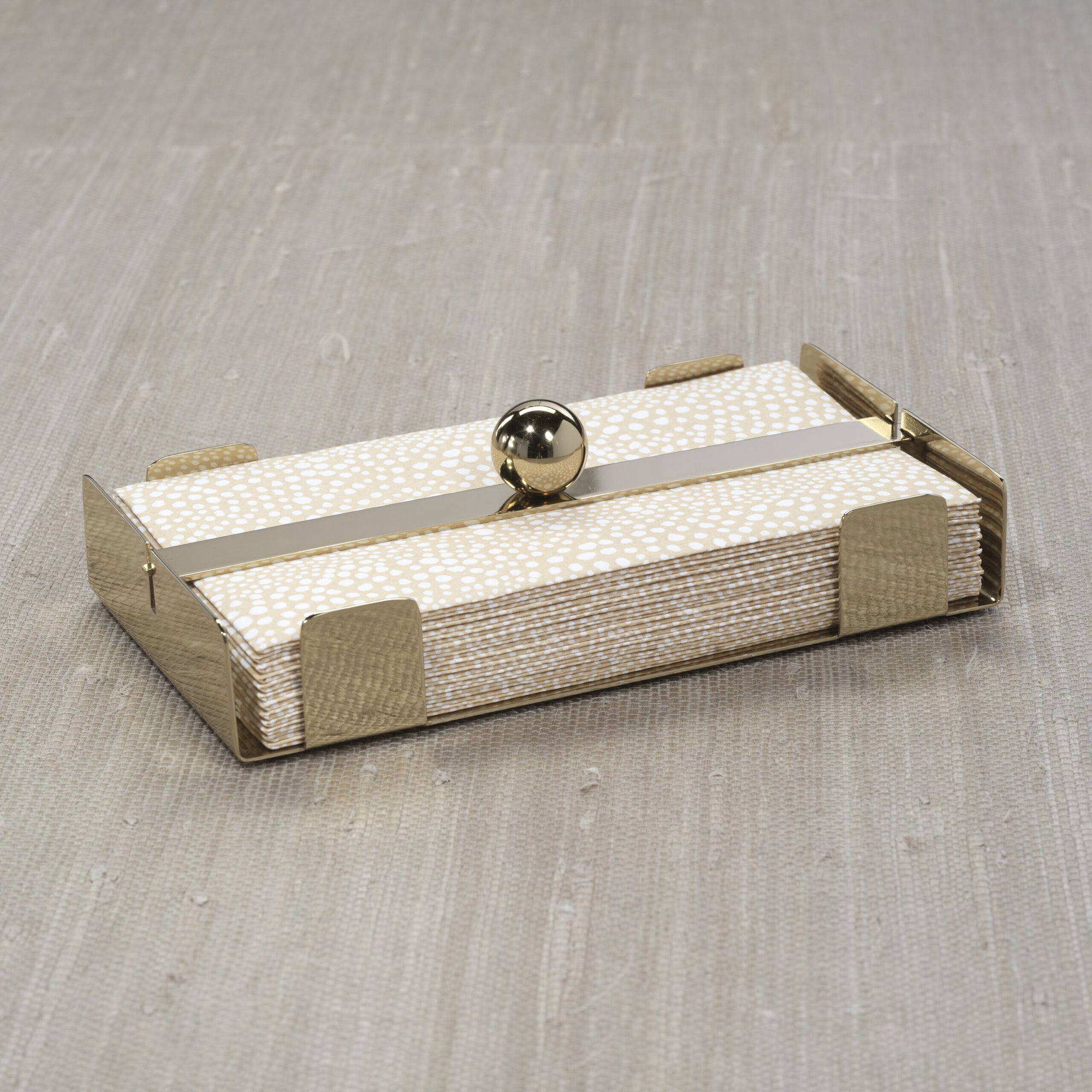 The Zodax Dinner Napkin Holder Gold displays a stack of white napkins with gold dots. This chic bar accessory, featuring a round gold ball on top, sits stylishly on a light wood surface.