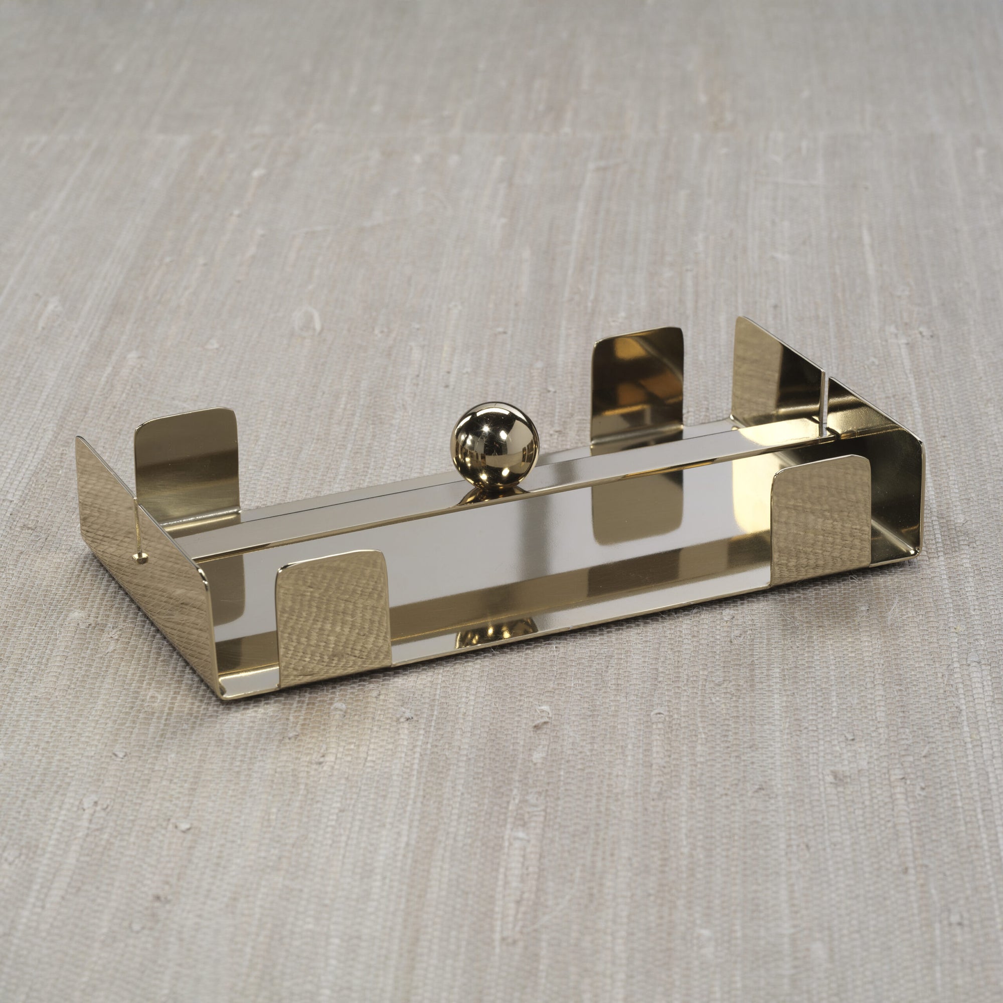 The Zodax Dinner Napkin Holder Gold is a rectangular steel napkin holder with four upright edges and a central round ball, making it a stylish bar accessory, shown here on a textured beige surface.
