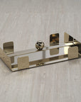 The Zodax Dinner Napkin Holder Gold is a rectangular steel napkin holder with four upright edges and a central round ball, making it a stylish bar accessory, shown here on a textured beige surface.