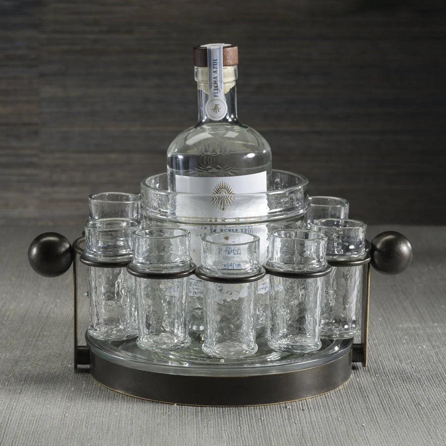 The Zodax Ten Shot Tequila Set w/Ice Bucket features a tall bottle at the center, surrounded by eight textured shot glasses, all arranged on a gray surface with a striped backdrop.