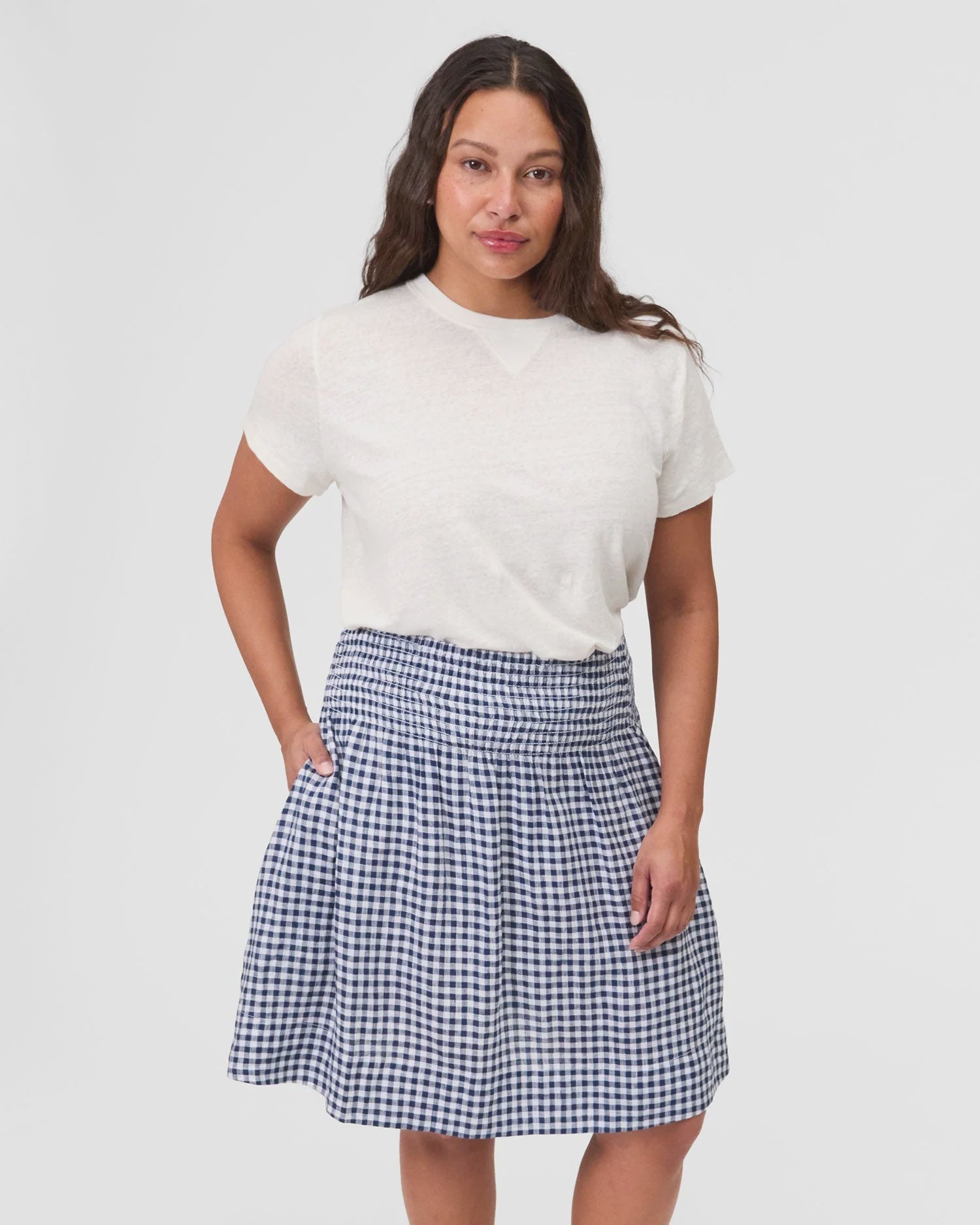 A woman with long brown hair models the Clare Vivier Zoe Skirt—a blue-and-white checkered skirt with a smocked waist—paired with a white short-sleeve shirt, standing against a plain light background.