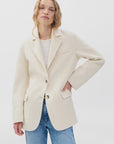 A person with wavy blonde hair models the Rag & Bone zoe wool blazer in cream over a white top and blue jeans, standing against a plain white background. One hand is on their hip while the other hangs at their side.