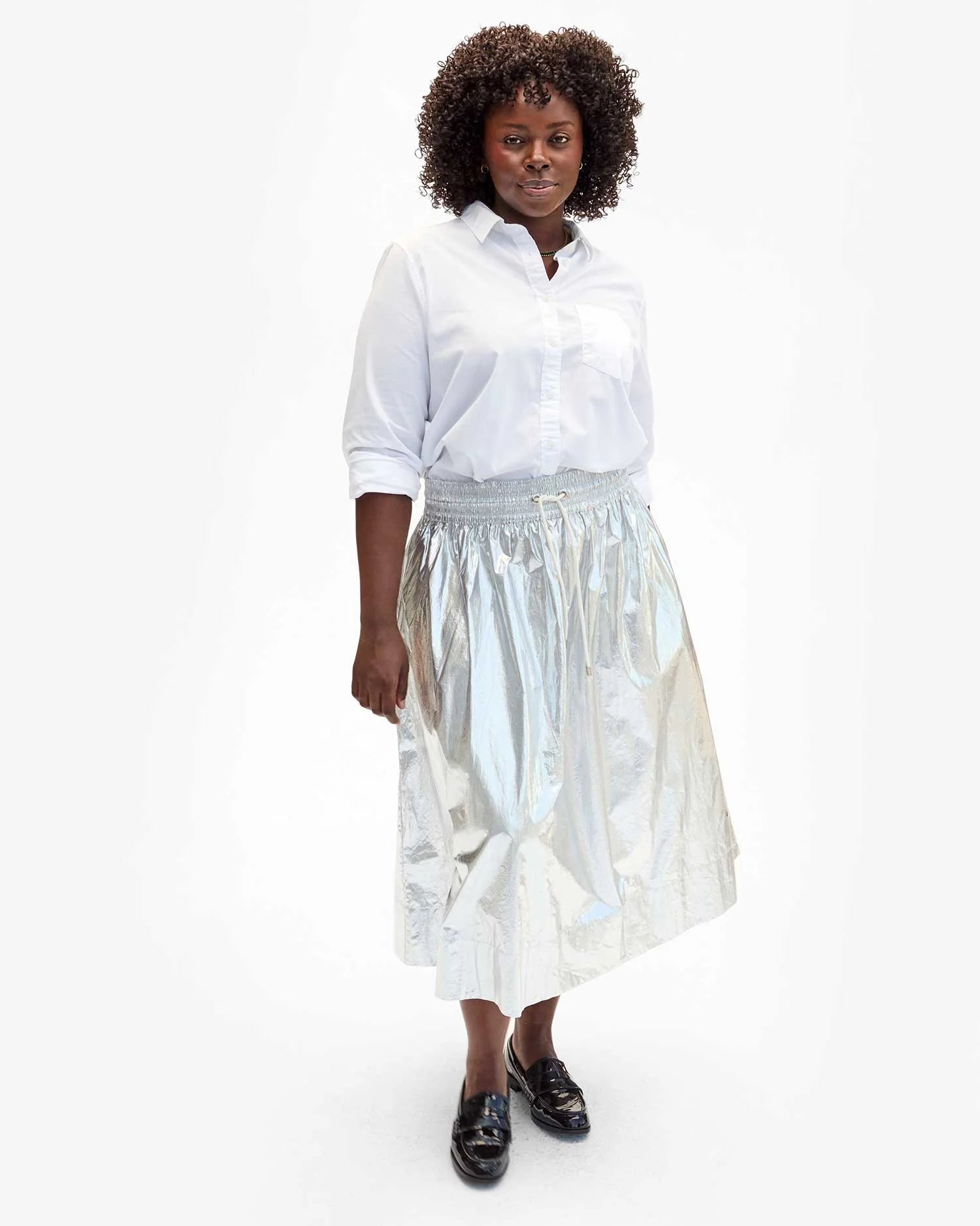 A person stands confidently in a white button-up shirt and the Clare Vivier Zoelle Skirt, which features a metallic smocked waist with a cotton metallic coating. Their curly hair complements the sleek look, enhanced by black loafers. The simple backdrop highlights this fashion-forward ensemble with its convenient side seam pockets.