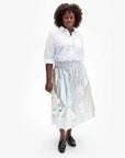 A person stands confidently in a white button-up shirt and the Clare Vivier Zoelle Skirt, which features a metallic smocked waist with a cotton metallic coating. Their curly hair complements the sleek look, enhanced by black loafers. The simple backdrop highlights this fashion-forward ensemble with its convenient side seam pockets.