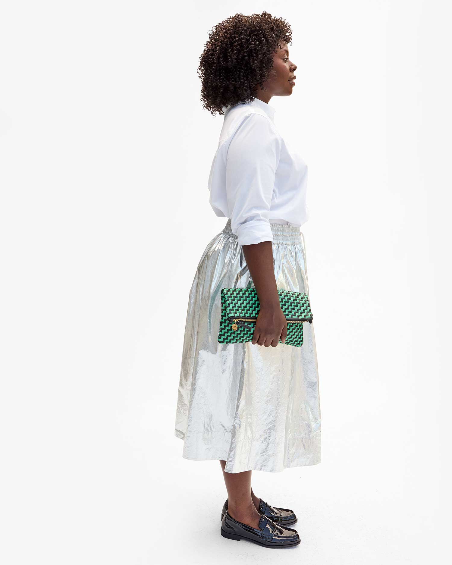 A person stands in profile against a white background, showcasing a white blouse, the Zoelle Skirt, and black shoes. They are holding a green and black checkered clutch from Clare Vivier.