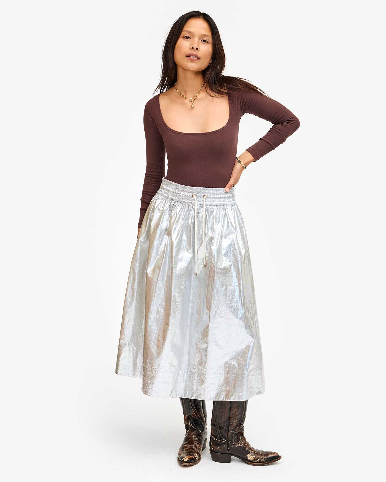 A person poses against a white background, exuding confidence with their long hair flowing as they confidently wear a brown long-sleeve top paired with the metallic Zoelle Skirt by Clare Vivier and patterned knee-high boots.