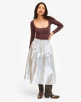 A person poses against a white background, exuding confidence with their long hair flowing as they confidently wear a brown long-sleeve top paired with the metallic Zoelle Skirt by Clare Vivier and patterned knee-high boots.