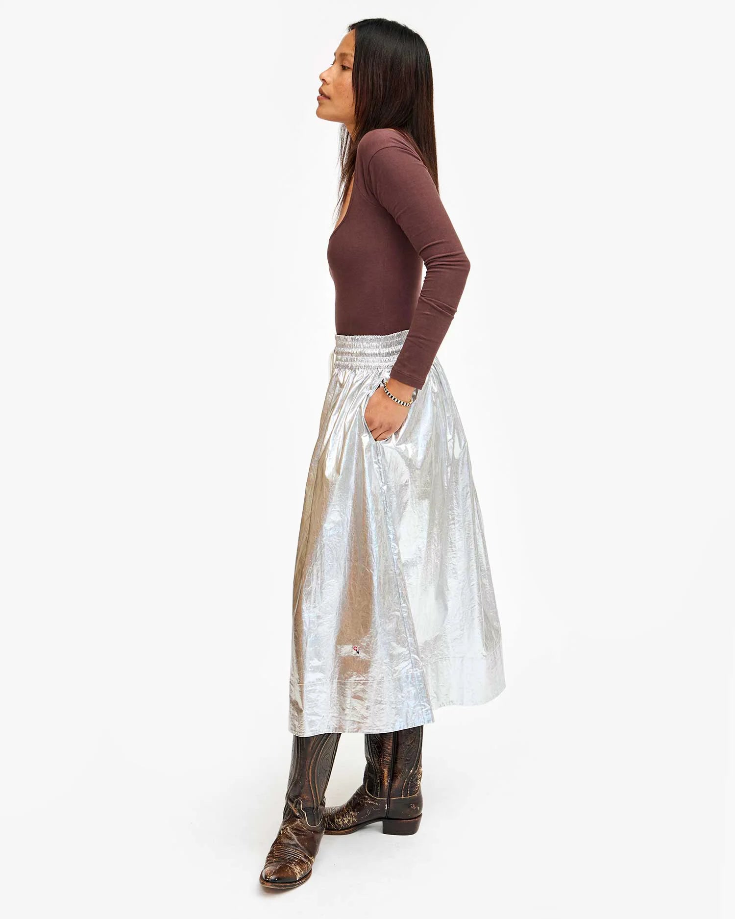 A person stands sideways wearing a long-sleeved brown top and the "Zoelle Skirt" by Clare Vivier, featuring a cotton metallic-coated fabric and an elegant smocked waist. Their patterned brown boots perfectly complement the ensemble, while their hands rest in the side seam pockets. They have long dark hair cascading against a plain white background.